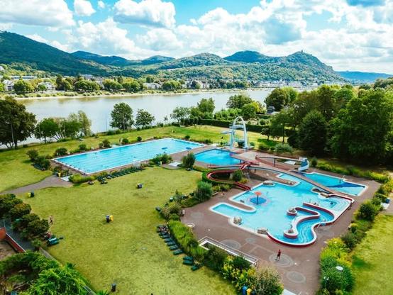 Freibad Rüngsdorf mit verschiedenen Becken und Rutschen, umgeben von grünen Wiesen und Bäumen, mit dem Rhein und Siebengebirge im Hintergrund.