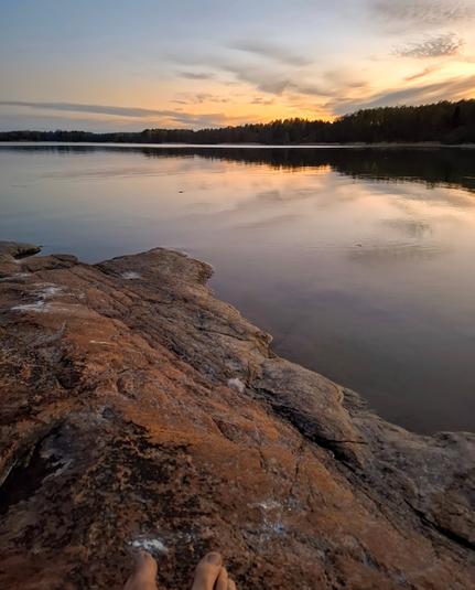 Sunset over a still inlet of the Guild of Finland with a forested shore a few hundred meters away and someone's toes on a sloping reddish granite shore in the foreground.