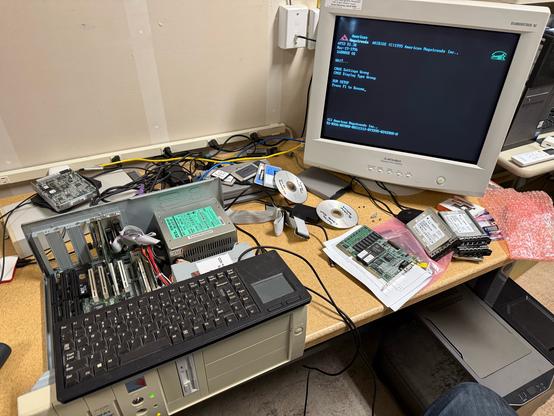 A somewhat cluttered workbench with an old baby AT style case in beige. A black 65% mini keyboard rests on the open case. The motherboard is partially visible in the case. Behind, we can see a large old CRT monitor, some CDs some peripheral cards and some more modern hardware. The CRT is showing the boot screen for a Pentium 100 BIOS