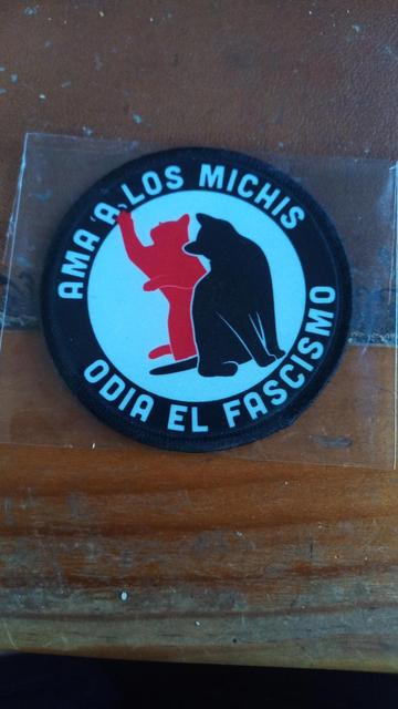 Es un parche para ropa redondo. En el centro dos gatos, uno rojo y otro negro en un círculo blanco. El gato rojo con la pata mueve una letra A que está en la leyenda que hay alrededor del círculo blanco y que reza: ama a los michis odia el fascismo. El parche está metido en su bolsita de celofán y apoyado en una mesa de madera y cuero.