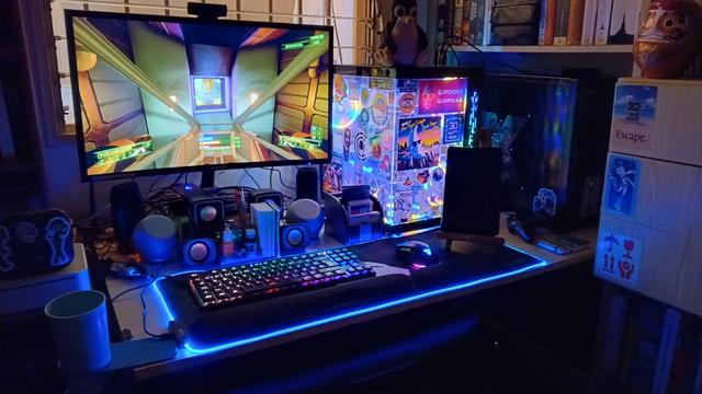 A photo of a gaming PC running the game Overload. There is plenty of RGB all around and a Tux plushie sits atop the case.
