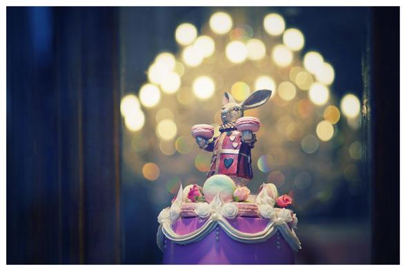 In einem Schaufenster steht auf einer mehrstöckigen Torte ein rosa Kaninchen, es hat zwei Macarons in den Händen. Der Kronleuchter, der auch im Hintergrund befindet, ist durch die offene Blende nur als eine Ansammlung von Lichtflecken zu erkennen.