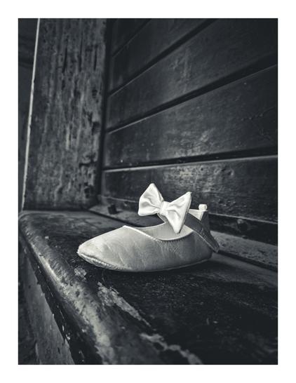 The photo is a black and white photograph of a single baby shoe. It is specifically a light-colored, satin-like shoe with a white bow. It is near the centre of the photo, toe side facing to the left. It is on a dark, weathered stone step. Behind the shoe you can see the lines from the dark shop shutter.