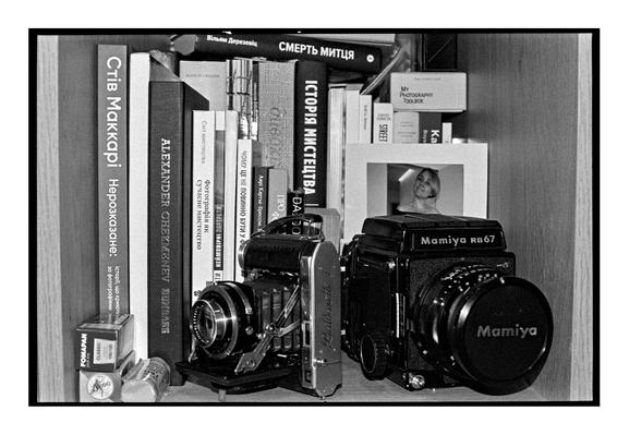 A black-and-white photograph of a bookshelf featuring two vintage cameras—an Agfa Isolette folding camera and a Mamiya RB67 medium format camera. They are surrounded by photography and art books. A small portrait of a woman is displayed between the books and cameras. The scene evokes a nostalgic and artistic atmosphere, blending analog photography with intellectual pursuits