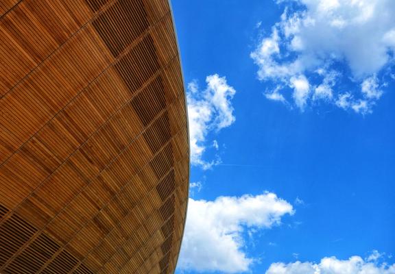 The image shows a section of the Lee Valley VeloPark in London, specifically the Hopkins Velodrome. A curved exterior wall, constructed from horizontally-laid wooden slats, dominates the left side of the frame. The wood has a warm, orange-brown tone. The right side of the image reveals a vibrant blue sky dotted with fluffy white clouds. The perspective is from a low angle, emphasizing the height and curvature of the building.