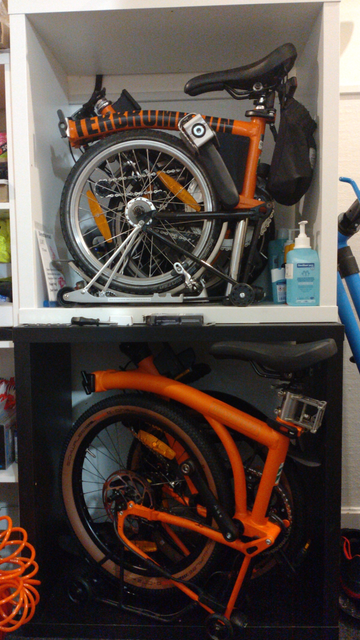 Zwei IKEA Kallax-2x2-Regale aufeinander. Oben ist ein klassisches 16" Brompton gefaltet darin. Unten passt ein gefaltetes Brompton G-Line so grade nicht hinein. Beide Räder sind orange, das obere Kallax ist weiß, das untere schwarz.