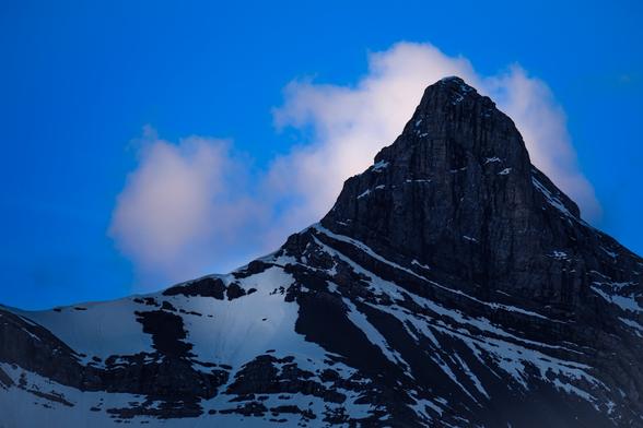A close up portrait of a single peak of Canmore Alberta's 
"Three Sisters" group of montains against a deep blue sky with a white cloud just behind the summit. Early morning sunlight is highlighting the left side of the cloud, and reflecting on a patch of snow below ridge.