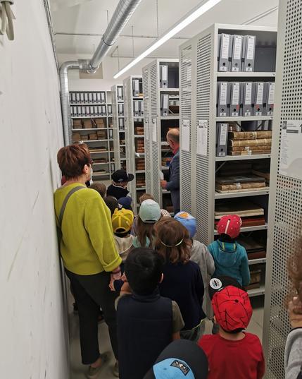 Die Kinder und ihre Erzieherinnen erkunden die Archivräume. Kindergartenkinder mit Erzieherin in einem Gang neben Regelreihen mit Akten und Büchern.