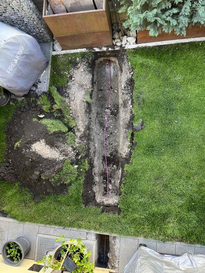 An overhead view of a garden showing a freshly dug trench with loose soil. A small pink fiber-tube runs through the trench.