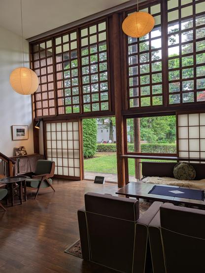 Inside of a mondernist Japanese living room built in 1940, during WW2. Two storey windows with a wood lattice open to a garden. Inside, wood floors, and a squareish couch in front of a wooden table.