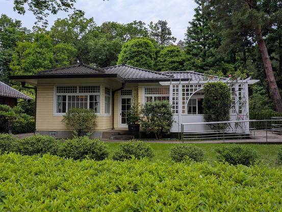 Exterior view of a 1920s Japanese house built with strong European influences. One story, with a simple gabled roof made of tile. The walls are pale yellow slats with white framed windows. A trellis covers a porch on the side