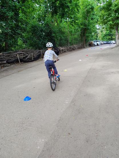 Das Bild zeigt ein Kind mit weißem Helm, hellblau gemustertem Pullover und blauer Hose von hinten, welches auf einem roten Fahrrad auf einem Weg um bunte kleine Hütchen Slalom fährt.
