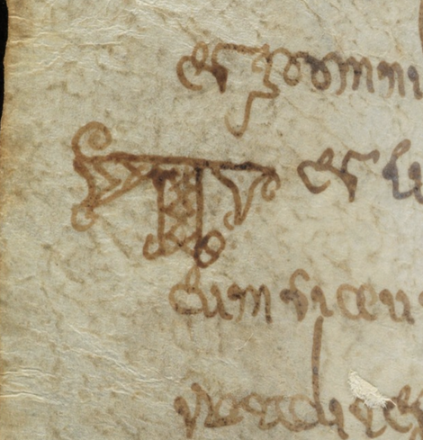Handwritten capital initial T surrounded by other text, all in brown ink. The T has zizag decoration and looks like it has two little feet running forwards.