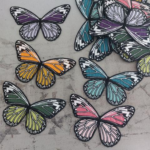 Sur un tissu gris tâché par de la colle textile (c'est une table à repasser) se trouvent 4 papillons en coton brodé dans des nuances de : violet, orange, vert, bleu et rose.
Dans le coin supérieur droit, on distingue une pile de papillons en tissus brodés dans ces mêmes coloris.