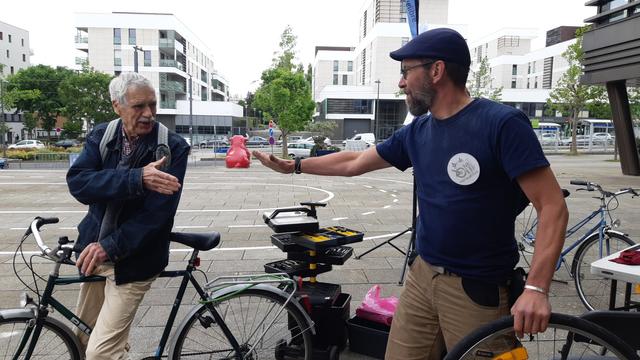 2 hommes (un ancien à vélo et un tenant une roue à la main) se saluent devant une servante à outils et des vélos, place de l'Europe, Hérouville Saint-Clair