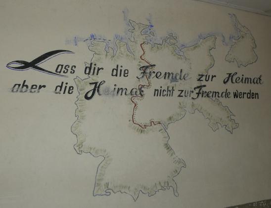 Eine Wand in einer Baracke:
Gemalte Europakarte.
Darüber ein  text:
"Lass dir die Fremde zur Heimat,
aber die Heimat nicht zu Fremde werden."