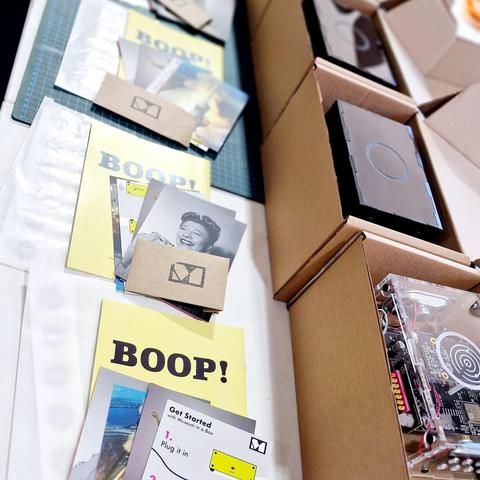 Visible are 3 Museums in a Box sitting quietly in small carboard boxes. The lids of the boxes are open to show the Museums in a Box sitting snuggly, before the boxes are a series of postcards and a booklet with Boop on the front. The postcards that are visible show the Apollo 11 launch, Billy Holiday and a Get Started with MiaB card.