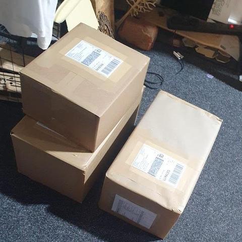 3 stacked labeled boxes awaiting pick up in DoES Liverpool. In the background can be seen part of a wing from a small plane and small laser cut wooden trees on a blue carpet.