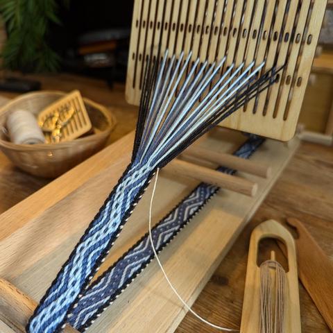 Photo d'un métier à tisser type inkle loom en bois, avec un galon en cours de tissage dessus (motif en zigzag en fil de coton bleu foncé et bleu clair, avec des bordures noire tissé au peigne d'encroix)