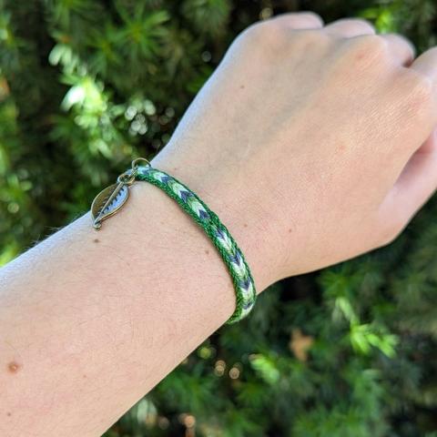 Photo d'un bracelet tissé porté au poignet avec une petite feuille en laiton. Fils de coton fins tissés dans un motif de chevrons mêlant des douces teintes de vert, blanc et bleu-gris, rappelant les couleurs d'une éclaircie dans un jardin après la pluie.