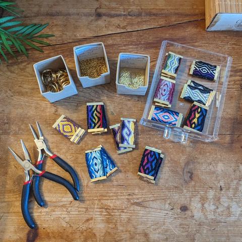 Photo d'une table en bois couverte d'un ensemble de pièces tissées aux couleurs de  drapeaux queer, et de matériel et apprêts dorés attendant d'être assemblés en bijoux tissés.