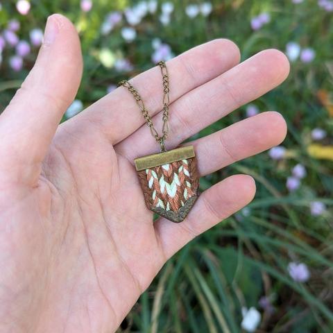 Photo d'un pendentif tissé tenu dans une main. Fine chaine et embouts en laiton encadrants un écusson de fils de coton tissés dans un motif en chevrons mêlant des couleurs de feuilles automnales : brun, caramel, vert clair et pointes de blanc pour rehausser le tout.