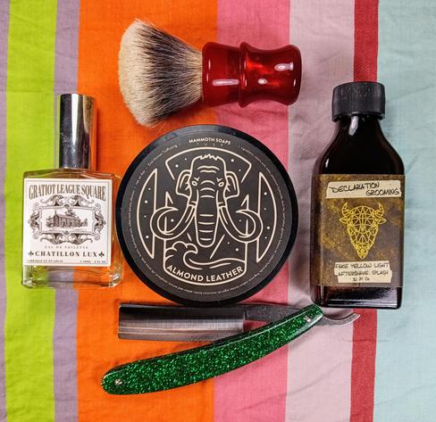 (Le Chat Mistral description)

The image showcases a collection of grooming items arranged on a striped background. At the top left, there's a bottle of "Gratiot League Square" cologne by Chatillon Lux. Next to it, a shaving brush with a red handle is placed above a round tin of "Mammoth Soaps" almond leather shaving soap. To the right, there's a small bottle labelled "Declaration Grooming" with a yellow geometric design, containing "Fake Yellow Light" aftershave splash. At the bottom, a straight razor with a green glittery handle is displayed. The items are neatly organised, creating a visually appealing setup.