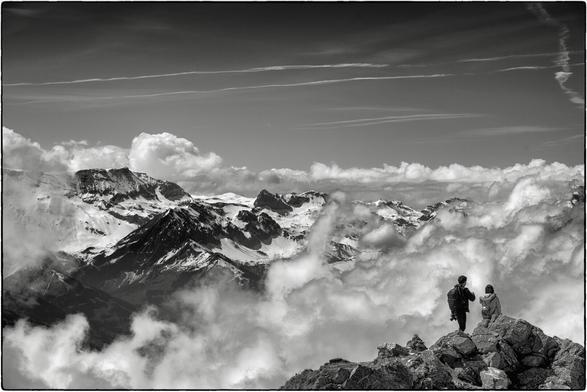 Schwarzweissfotografie eines Bergpanoramas mit Wolken. Im Vordergrund (rechte untere Ecke) sind 2 Personen zu erkennen.