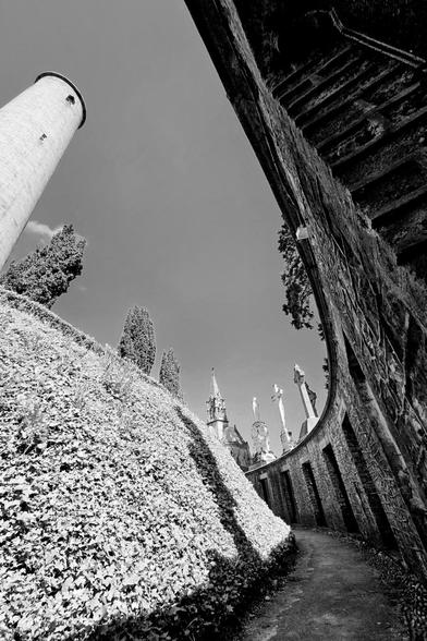A low-angle, black and white photograph shows a tall, cylindrical stone tower on the left and a curved brick structure containing doorways to tombs on the right, forming a unique architectural composition within Glasnevin Cemetery. The sky is visible in the open space between the two structures. The ground slopes upwards towards the right, covered in dense foliage, with several religious statues and a church spire visible in the background. A narrow curved pathway beside the tombs on the right.