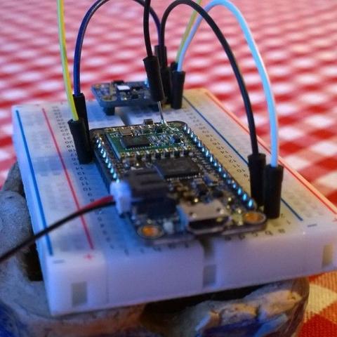 The picture shows a small white electronic device connected to multiple wires, which are hooked into another board labeled 'Arduino Uno'. The Arduino is on a breadboard with various components. There is also a microcontroller board visible, suggesting the setup might be for a DIY electronics project or hobbyist activity. The scene is casual and appears to be set in a domestic environment, possibly a kitchen counter due to the presence of what looks like wooden elements and a fabric that could be a tablecloth.