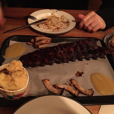 "This picture shows a table with a platter of BBQ ribs, mashed potatoes, and honey mustard sauce, ready to be enjoyed. The table also holds a cupcake. In the background, there is a person sitting at another table with a plate of food."