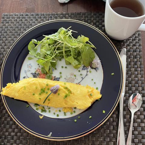 A plated French omelette garnished with chopped chives and flower, served alongside a small salad of arugula. A cup of tea is placed next to the plate on a woven placemat