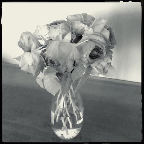 This picture shows a clear glass vase holding an arrangement of white flowers with green leaves on a table. The flowers are arranged in a somewhat casual manner, and the vase is placed in front of a light-colored surface which appears to be a table or shelf. There is no visible text within the image.