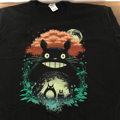 "A black t-shirt featuring a graphic design of an anime-style character, resembling a cartoon cat, standing on rocky terrain surrounded by lush greenery and a large tree. In the background, there is a full moon and a constellation. The cat's eyes are open and it appears to be looking directly at the viewer. The overall mood of the image is whimsical and magical."
