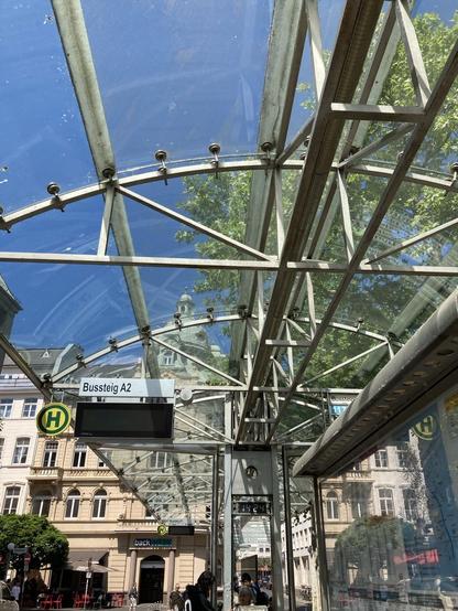 Zu sehen ist ein Foto, das in der Perspektive schräg nach oben fotografiert ist. Der Himmel leuchtet blau in den Facetten des Metall-Glasdachs der Bushaltestelle am Friedensplatz. Im Hintergrund Altbaustadthäuser. Rechts Baumgrün.