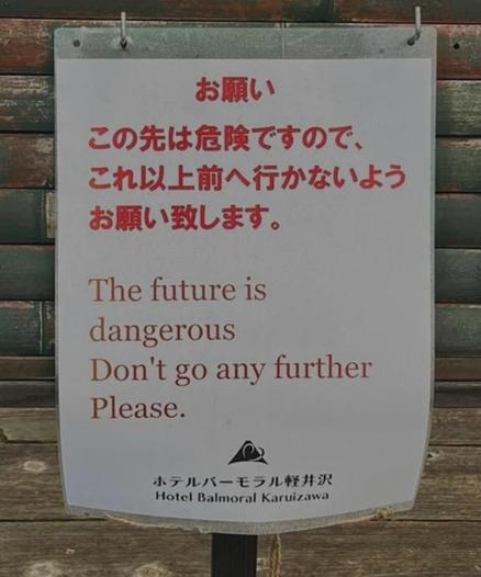 The image shows a sign with text in both Japanese and English. The sign is white with red and brown text. The Japanese text at the top reads "お願い この先は危険ですので、これ以上前へ行かないようお願い致します。" which translates to "Please, this area ahead is dangerous, so I kindly ask you not to go any further." Below this, in English, it seems to be an automatic translation to the message: "The future is dangerous. Don't go any further. Please." At the bottom of the sign, there is a logo and the text "ホテルバーモラル軽井沢" (Hotel Balmoral Karuizawa) in both Japanese and English. The sign is mounted on a wooden structure, possibly a fence or wall, with a metal hook at the top for hanging. The background is a wooden surface, likely part of a building or structure."