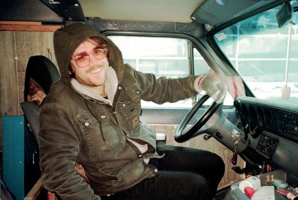a man in a winter coat and sunglasses sitting on the driver's side of a van, with a huge grin on his face, extending his right arm towards the passenger door i just opened