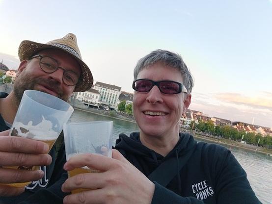 Zwei Personen stehen am Ufer eines Flusses und genießen ein Bier. Im Hintergrund ist der Rhein und ein Stadtteil mit Gebäuden und Bäumen zu sehen, und der Himmel ist in sanften Pastelltönen. Die beiden Personen lächeln und scheinen sich zu amüsieren.