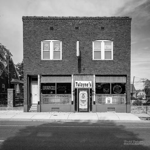 A black and white view of a two-story brick building.  The first floor has a sign saying "Tulayne's" which is the name of the bar.  The door is covered to look a Budweiser Natural beer can.  Beer related neon signs hang in the windows on either side.  A door at the left provides access for the upstairs portion of the building.