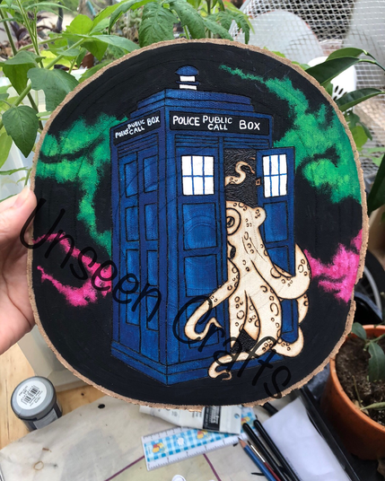 Oval wood slice with burned design showing an octopus coming out of a blue painted police call box with black background with vibrant green and pink streaks.
