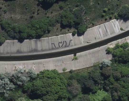Aerial photograph of a section of a creek that has been channelized with concrete banks. The creek flows from left to right across the photo and gently curves towards top of the photo. Around it is a scrub with low greenish plants and a few deciduous trees with leaves on.

The concrete banks of the creek are stained with water and dirt. There is some graffiti on the banks, and in particular, on the bank closer to top of the photo there is a large painted kaomoji "shruggie" (an arrangement of ASCII and kana symbols that looks like a person shrugging their arms).
