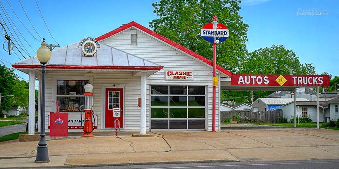View of a vintage Standard gas station with old signs, clock, gas pump, air hose, etc. 
The building is painted white with red trim.