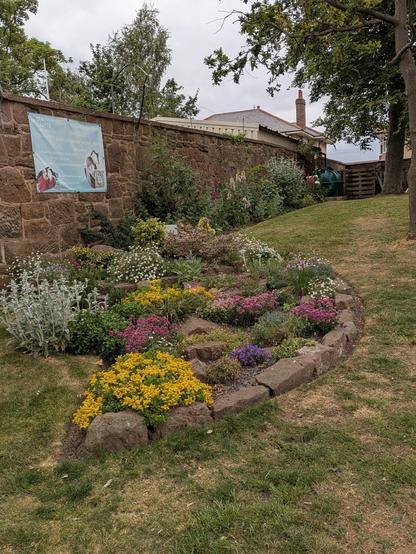 A beautifully landscaped garden features colorful flowers arranged in a stone-bordered bed next to a brick wall with a banner.