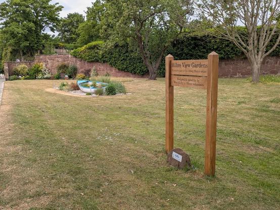 A grassy garden area features a wooden sign for Hilbre View Gardens and a small boat-shaped planter amidst trees and shrubs.