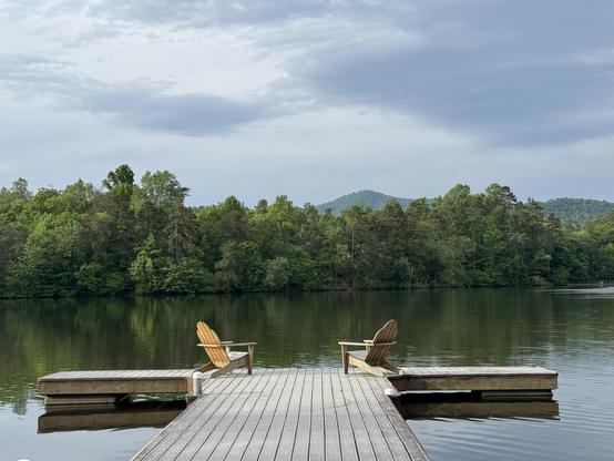 A calming photo of the community dock.  It is in the shame of a "T", jutting out into the very calm lake, with overcast skies.  The trees on the far bank are all fully leafed out, but in a handsome light green, while the mountains one can see behind them are also cloaked in green now.

Two Adirondack chairs are set on the top of the "T", partially facing each other, and the view of the lake.  Someday I'll have to do more than photograph these guys.

A large (?) frog barked at me as I was taking this picture, but he didn't show anything more than his disapproval of my presence.
