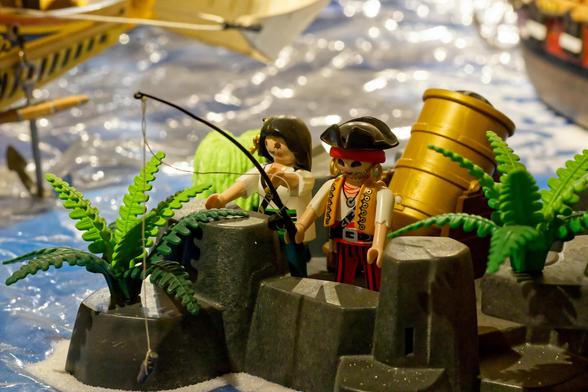 Two Playmobil pirate figures are positioned on a small island made of plastic rocks. The female pirate is holding a fishing rod, and it appears she has hooked a fish. The male pirate stands next to her, and a toy cannon is visible behind them. A pirate ship is visible in the background.
