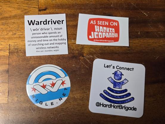 4 stickers sitting on a table. One is a meme definition of Wardriver, another is "As seen on Hacker Jeopardy," one is a WiGLE logo with the Chicago flag in the background, and the last is the Hard Hat Brigade logo sticker.