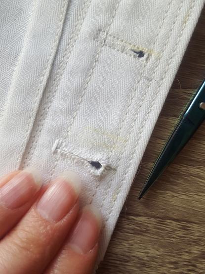 A very close up photo of a tailor's buttonhole sewn onto a white linen cotton twill fabric, the front edge of what is a corset but could not be recognised as such based on this photo alone. It is tiny, quite tidily done, but also with such coarse and thick, slightly fuzzy linen thread, that it looks more utilitarian than pretty. There's a bunch of different markings on the white fabric, as where the buttonholes wanted to go was up in the air for a while. There's multiple lines of reinforcement top stitching by machine, also. The stitches themselves are tiny and closely spaced. Sini's pink fingertips, oval natural nails, and the pointy end of thread snips in the frame.