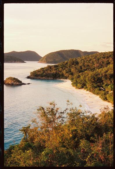 A film photograph of Trunk Bay, St John US Virgin Islands. The photo is taken from an overlook which offers a vantage point of the white sand beach from above. The beach is in a U-shaped cove and there is a small island close to shore at the far end of the beach. The water is turquoise. In the background you can see other points of land and islands. Tropical vegetation can be seen in the foreground.