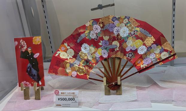 Ein Fächer steht aufgefaltet in einer Vitrine.
Es dominiert die Farbe rot.
Ein Preisschild zeigt 500000 Yen.
Links daneben das Bild eines in schwarz traditionell gekleideten Mannes mit diesem Fächer in der Hand.